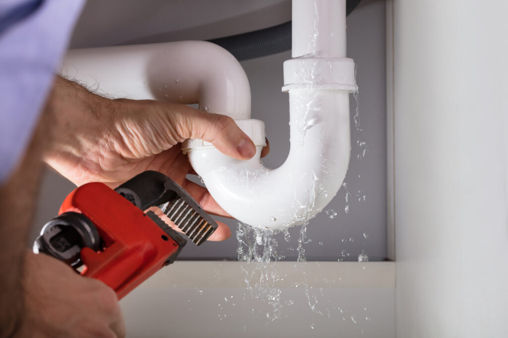 A man with a wrench working on a leaking drain pipe underneath a sink.