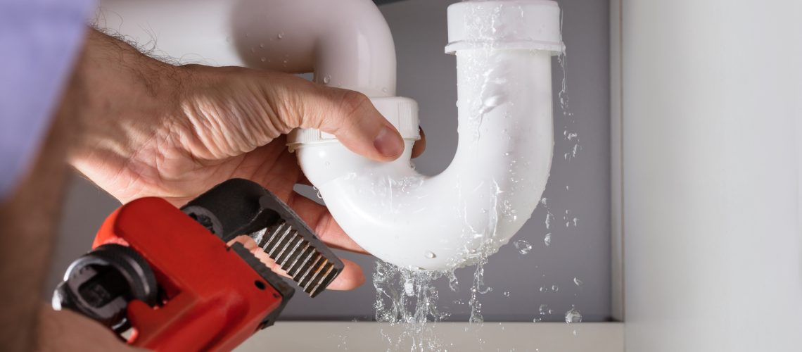 A man with a wrench working on a leaking drain pipe underneath a sink.